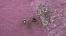 Two small metal hardware components and a pile of silver metal rings laid on a textured pink surface with some clear plastic wrapping visible.